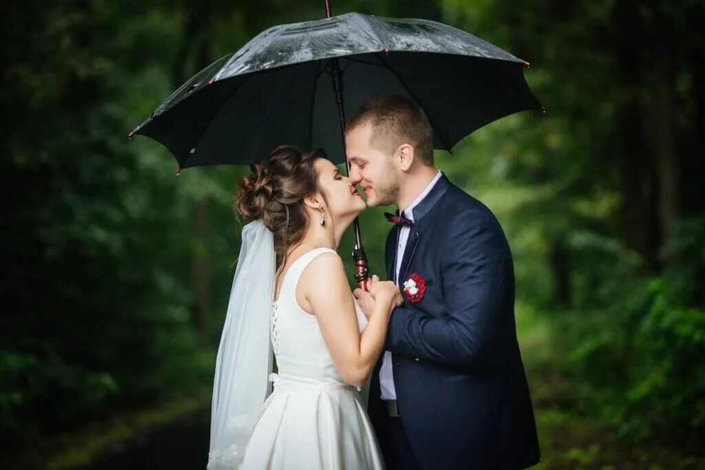 緑に囲まれて雨の中で傘をさしながらキスをする新郎新婦