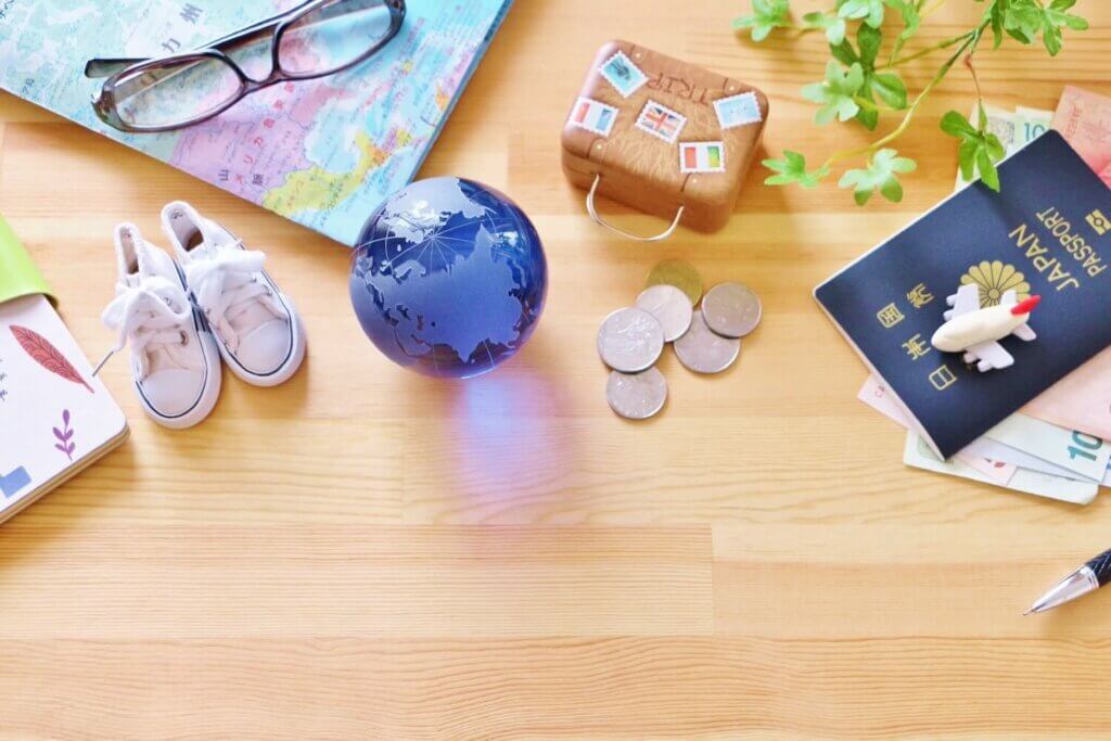 ミニチュアのガラス玉の地球と靴とカバン、パスポート、メガネ、地図が机に並べられている