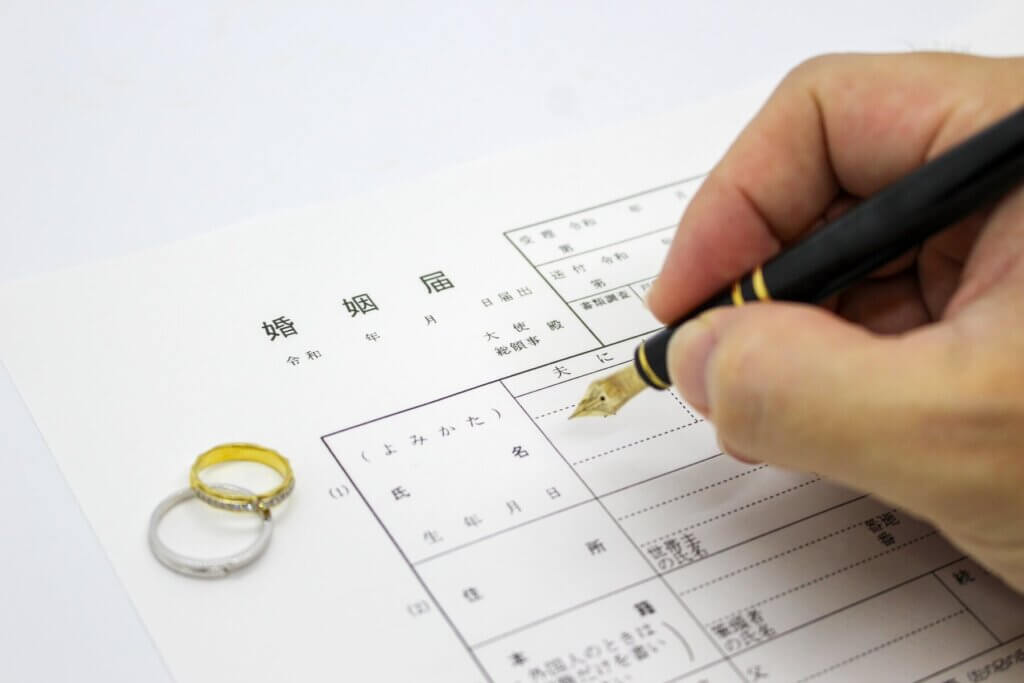 婚姻届に記入する男性ので。その届の上には1組の結婚指輪がのっている。