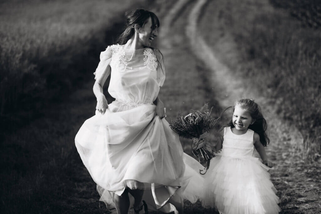 草原の中を笑顔で走る。フラワーガールの女の子と新婦のモノクロ写真