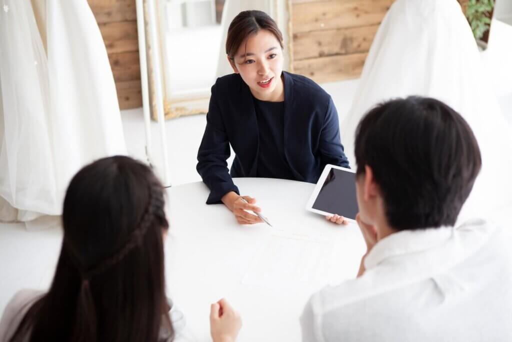 新郎新婦に向かい、左手にはタブレット、右手にはボールペンを持ち、笑顔で話をするウエディングプランナー
