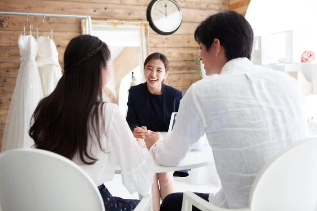 ウェディングドレスが釣られた衣装室で新郎と新婦に向かって笑っているウエディングプランナー