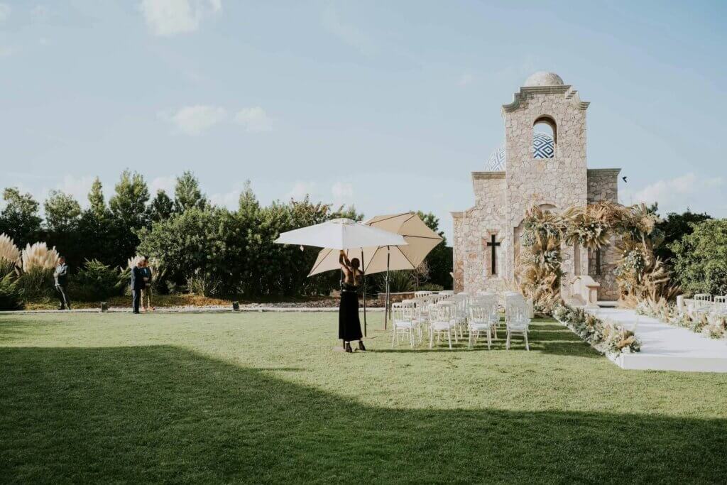 緑の森にたたずむ協会のガーデンに作られた白いバージンロードと白いケースと椅子そしてパラソルを開いている女性。