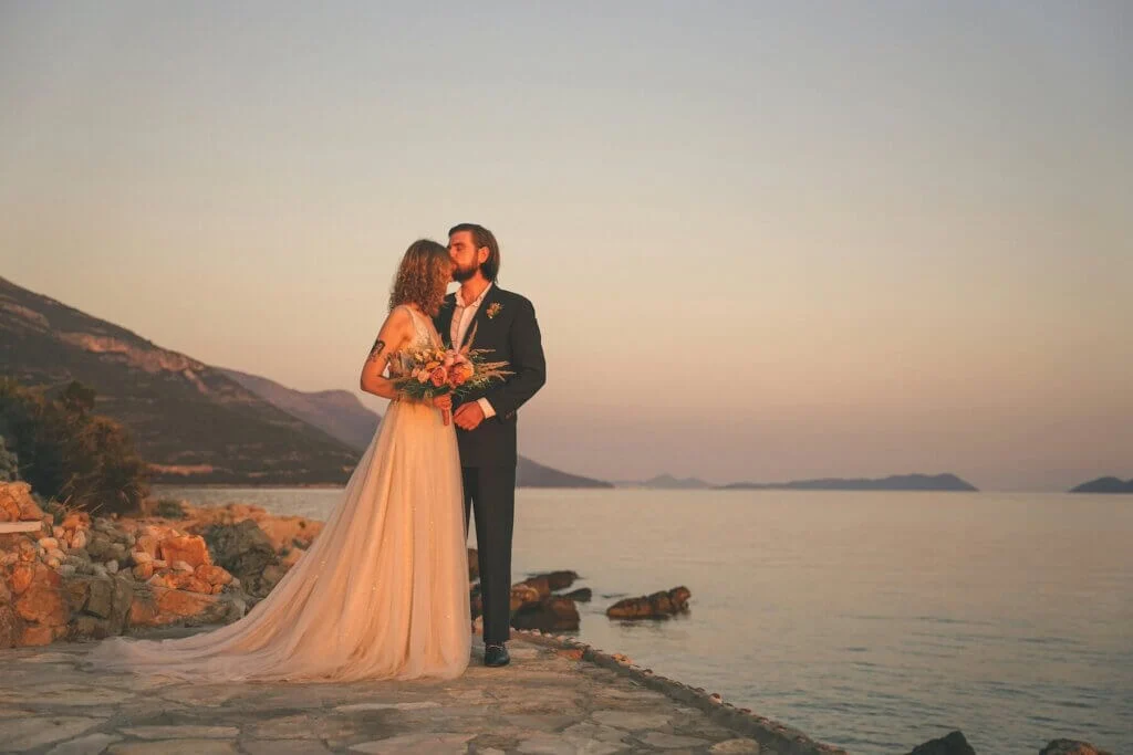 夕闇迫る海岸沿いで、タキシードとウェディングドレスに身を包み、新婦のおでこにキスをする新郎
