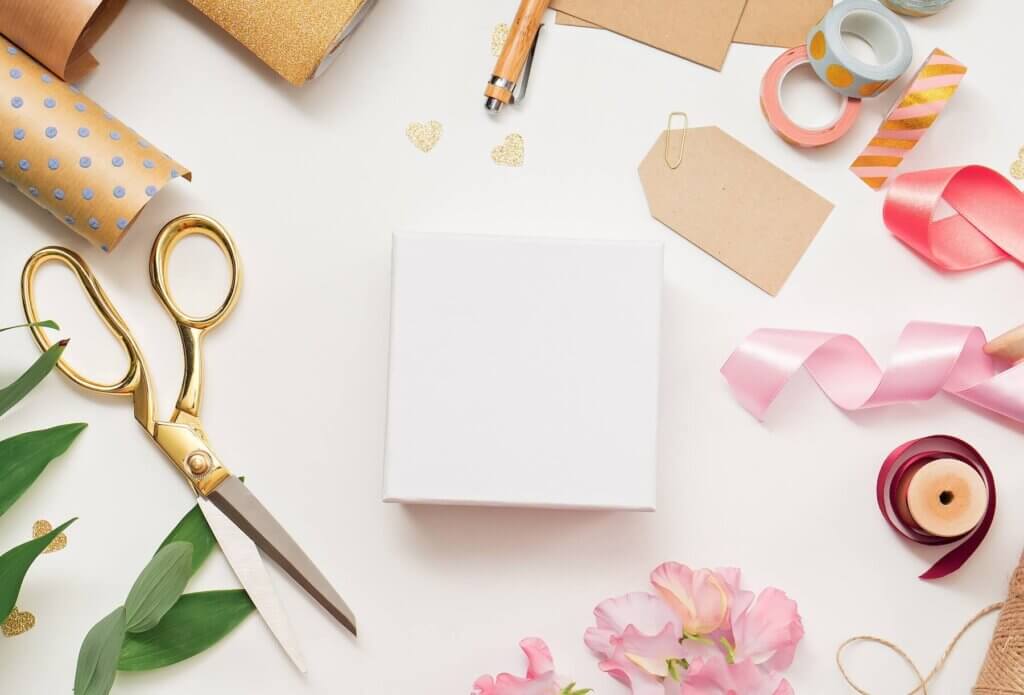 ナチュラル素材のクラフト紙とピンクや赤のリボン、大きなハサミ、グリーンの葉っぱなどのラッピング素材が並べられている