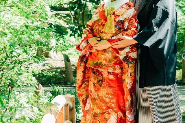 緑豊かな日本庭園で羽織・袴と色打掛で端の上で並んで立つ新郎と新婦。