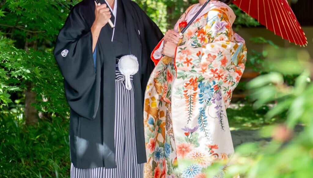 緑の森の中で番傘を持ち、向かい合って立つ羽織・袴の新郎と色家掛けの新婦