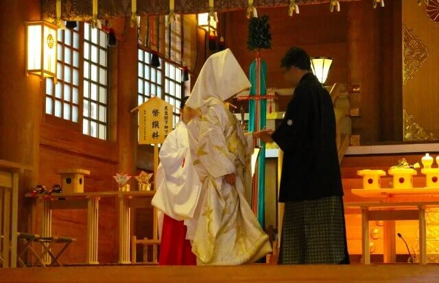 羽織、袴姿の新郎が白無垢姿の新婦の手を取りに、巫女さんのサポートを受けているシーン