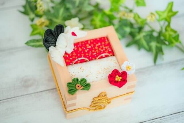 寿の文字と椿の花の飾り、そして鶴の飾りがあしらわれた枡の形をした結婚指輪のケースに1つの結婚指輪が収まっている