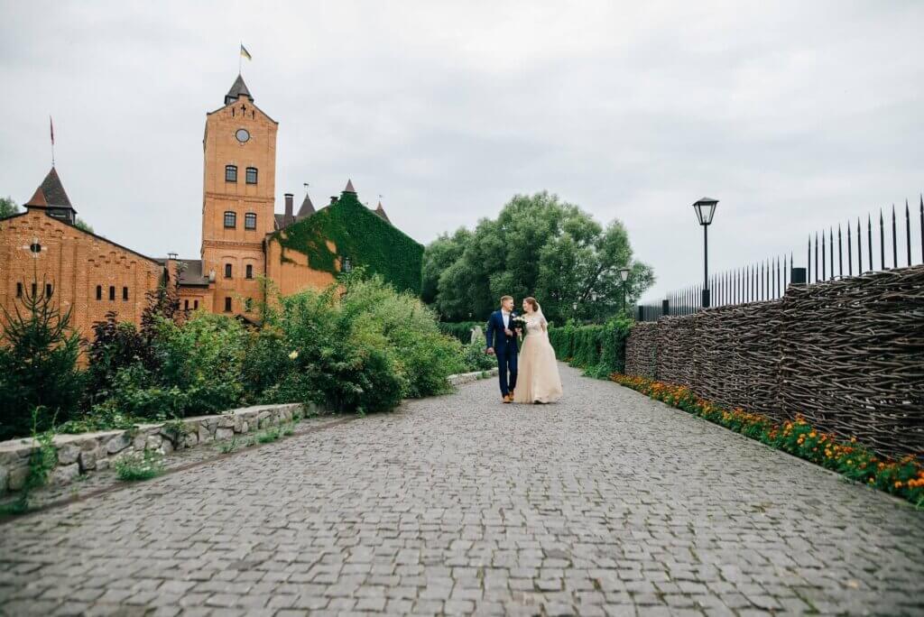 古いヨーロッパの石畳の街並みの中で腕を組み見つめ合って立つ、紺色のタキシードと薄いオレンジのウェディングドレスに身を包む新郎と新婦