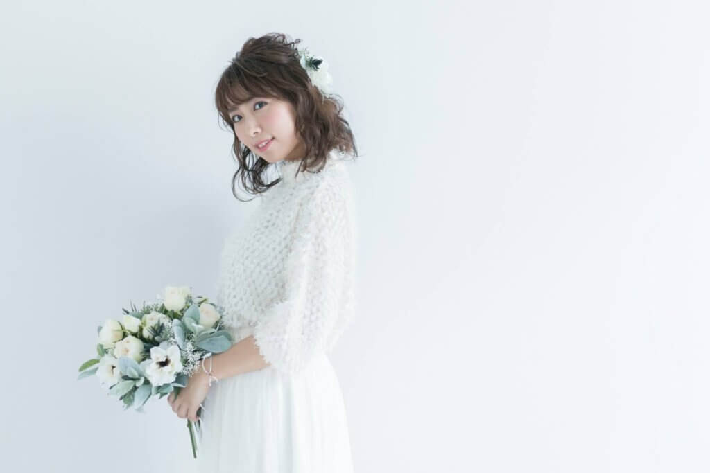 ロング袖でふんわりとしたイメージのウェディングドレスに身を包み、白いブーケを持ってこちらを向いて笑顔の新婦