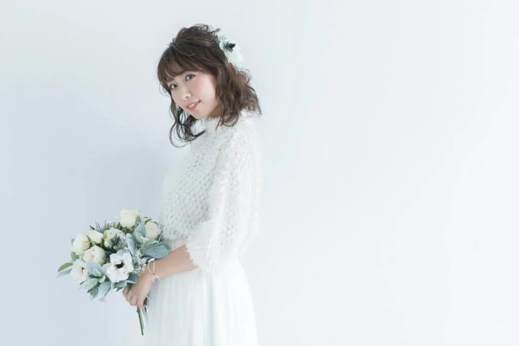 ロング袖でふんわりとしたイメージのウェディングドレスに身を包み、白いブーケを持ってこちらを向いて笑顔の新婦
