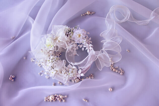 淡いパープルのレースの上に白い花飾りが置かれている。周りには真珠のアクセサリーが置かれている。