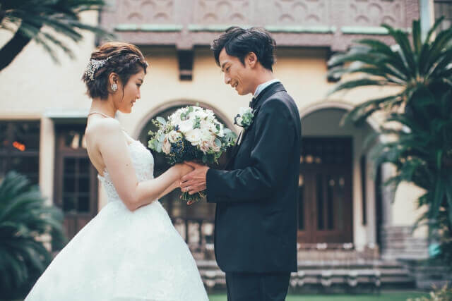 結婚式場のガーデンで、タキシード姿の新郎と、ウェディングドレス姿のブーケを、お互いの手でもって見つめ合っている