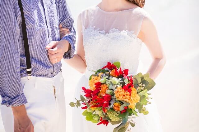 砂浜でカジュアルな服装の新郎と白いウェディングドレスにカラフルなブーケを持った新婦が腕を組んで立っている