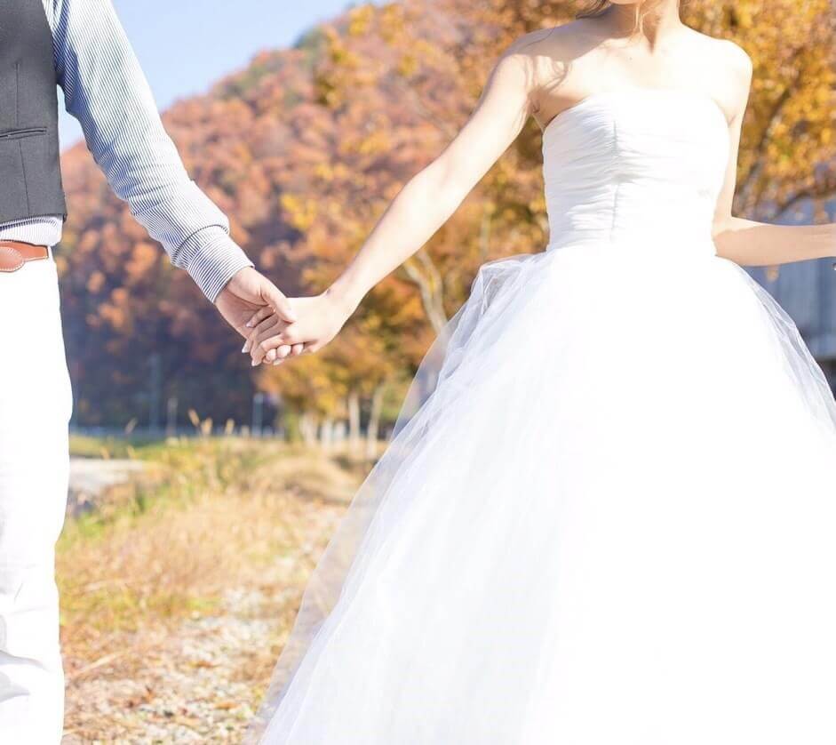 紅葉している山を背景に、カジュアルな服装の新郎と、白いウェディングドレスの新婦が手をつないでいる