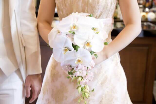 白い胡蝶蘭のブーケを持って、白いタキシードの新郎の横に立つ新婦