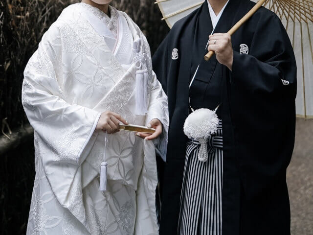 紋付袴と白無垢姿の新郎新婦。新郎が肩に番傘をもられかけさせている。