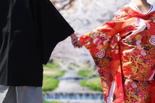 紋付袴姿の新郎と色打掛姿の新婦が桜と小川を背景に手を繋いでいる