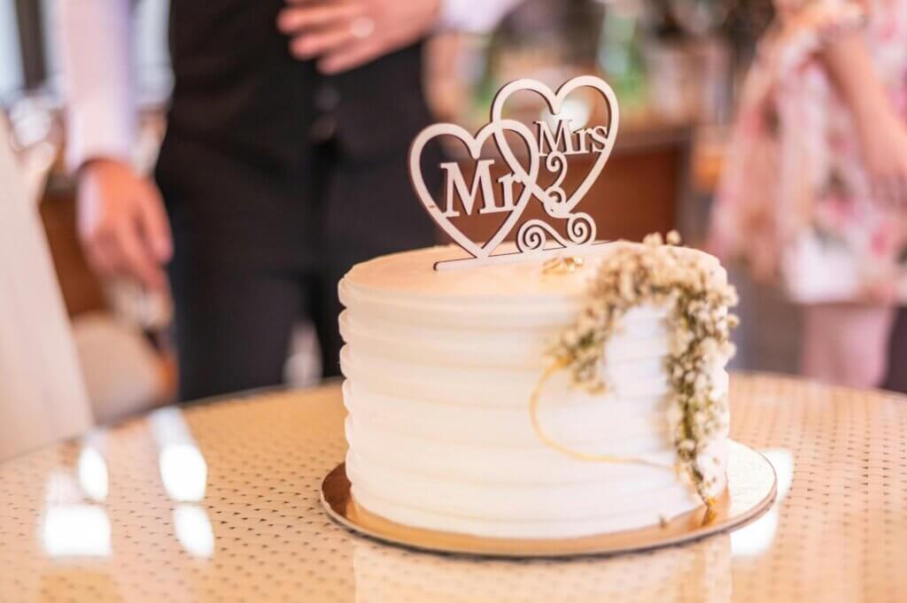 白いウエディングケーキの上に、MR、MRSの文字がハートの中に書かれているオブジェのっている。そのケーキの後に新郎新婦が立っている。