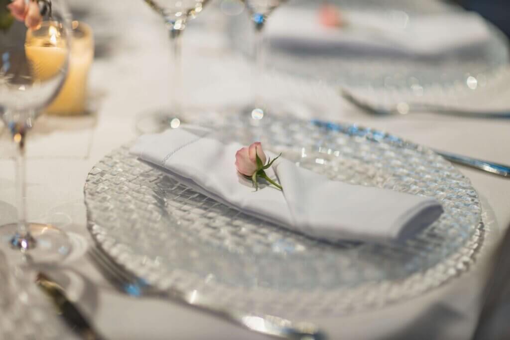 小さなピンク色のバラが白いナフキンに配置され、ガラスのお皿に上に置かれているテーブルコーディネート。