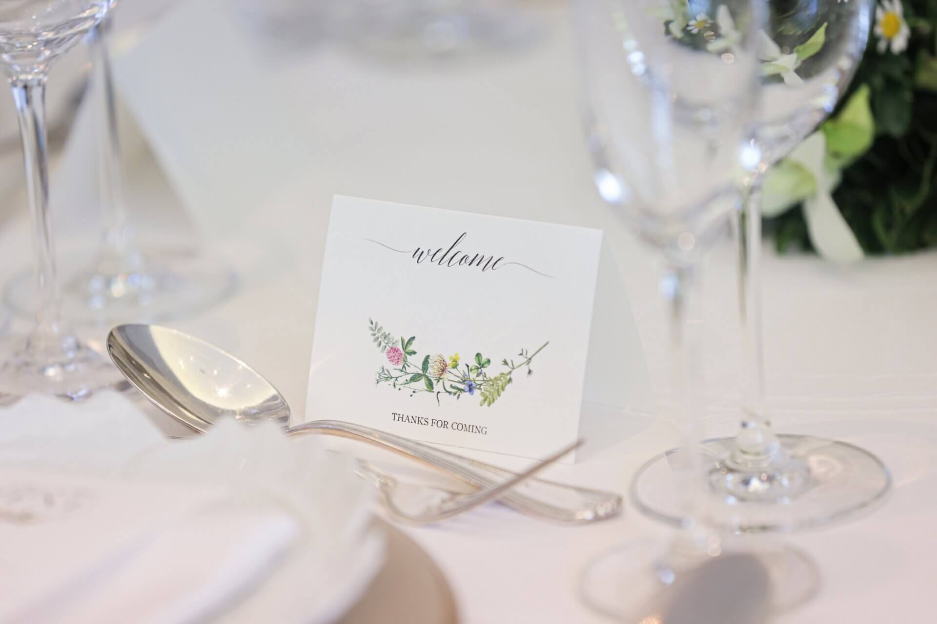 結婚式のテーブルに置かれた、ボタニカルなイラスト入りの白い席札とシルバーのカトラリー。
