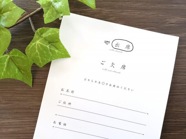 木目調のテーブルの上に置かれた、「御出席」「御欠席」の選択肢がある結婚式の返信ハガキとアイビーの葉