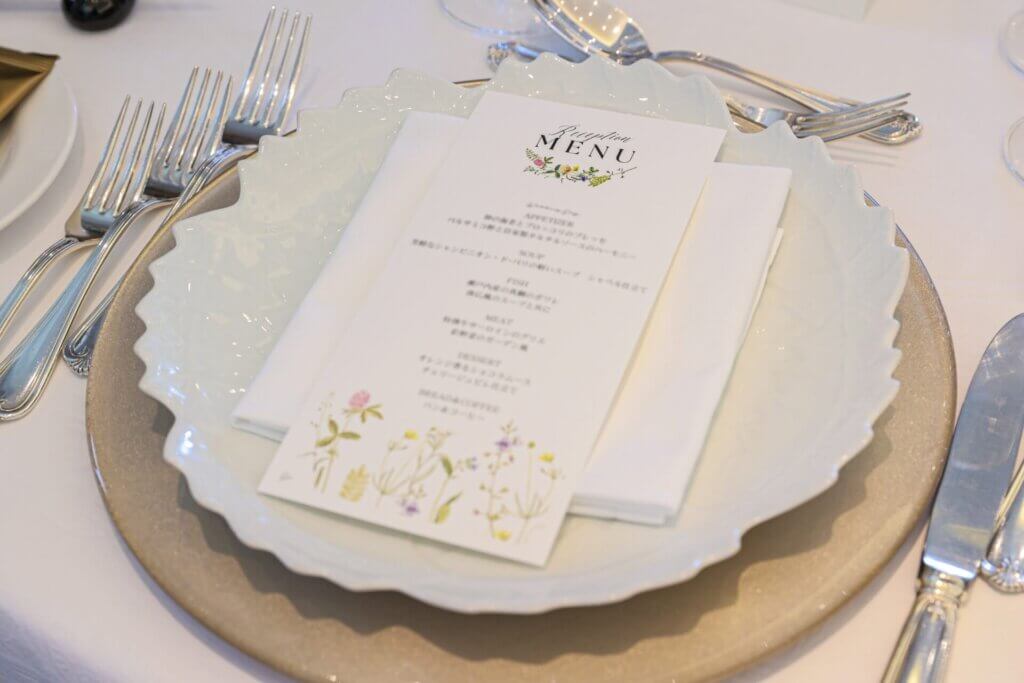 花のイラストを添えた結婚式のメニュー表。白いナプキンとフリル状の白い皿、カトラリーが並ぶ席次。