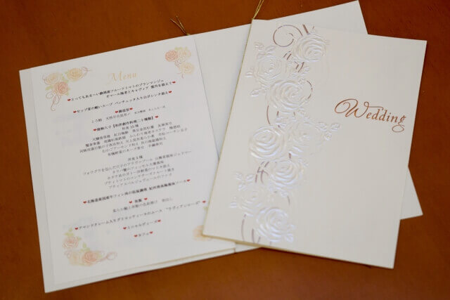 バラの型押しが施された白い結婚式メニューの表紙と、詳細な料理内容が記載されたメニューの中面。