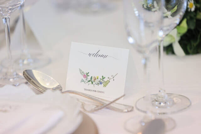 「welcome」と書かれた野花のイラスト入りのカード。披露宴会場のテーブルに置かれたグラスとスプーン。