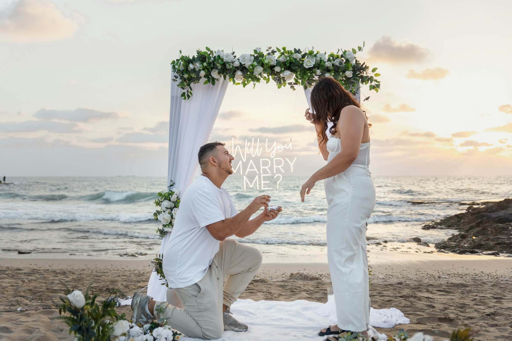 夕暮れの砂浜で、ひざまずいてプロポーズする男性と驚き喜ぶ女性の姿