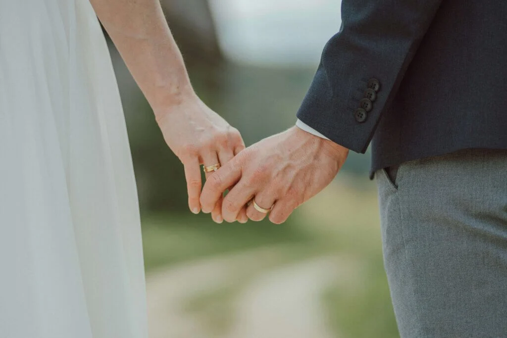 結婚指輪をはめた新郎新婦が、屋外で優しく手を繋いでいる手元のアップ