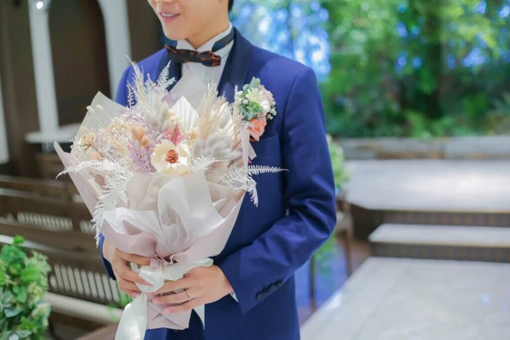 青いタキシードを着た新郎が、サプライズ用のドライフラワーの花束を持つ姿