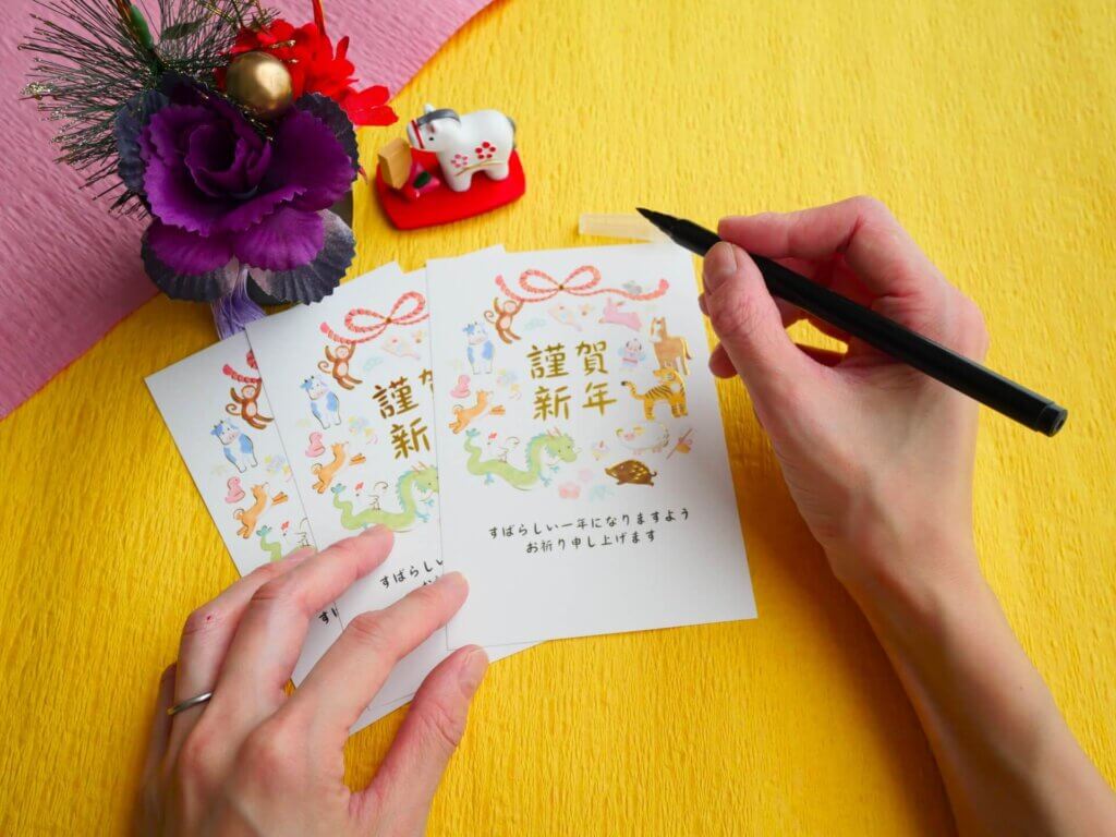 「謹賀新年」の文字と干支のイラストが描かれた、結婚報告を兼ねた年賀状に宛名を書く手元