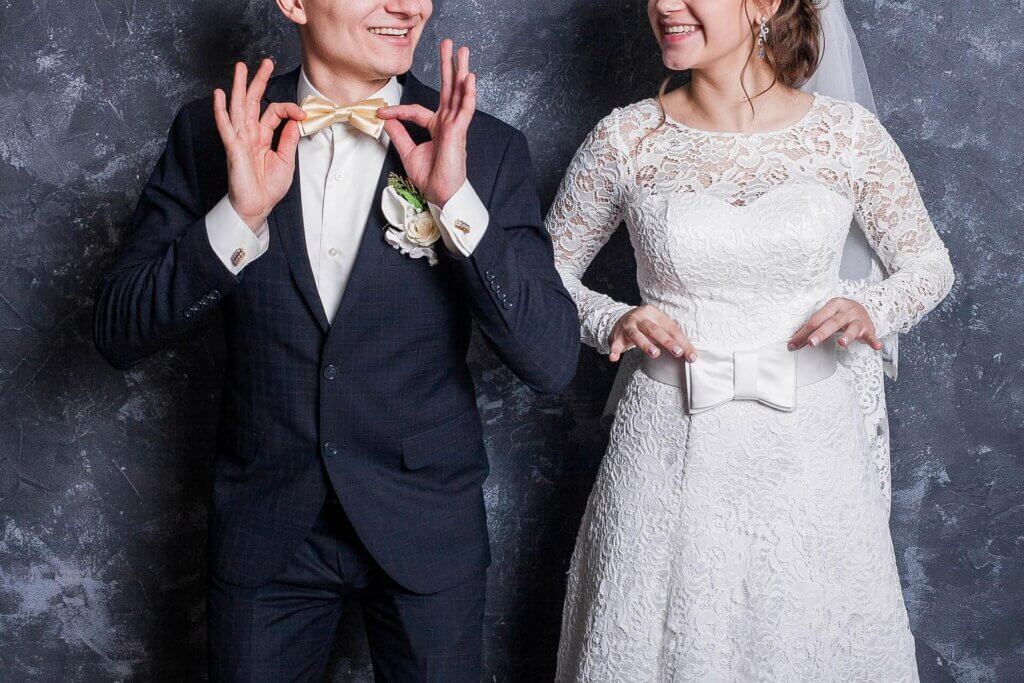 結婚式の新郎新婦。新郎は蝶ネクタイ、新婦はドレスのリボンに手を添えて笑顔。