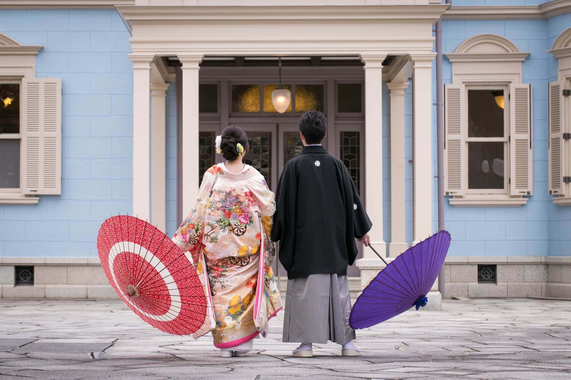 青い洋館の前で赤と紫の和傘を手にした和装の新郎新婦が立つ後ろ姿