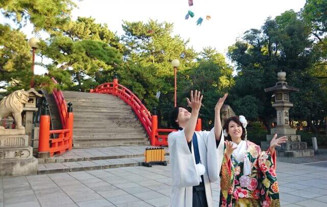神社の朱色の太鼓橋を背に折り鶴シャワーを楽しむ和装の新郎新婦