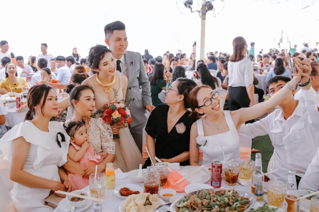 結婚式の披露宴会場で、笑顔の新郎新婦がゲストの友人たちと一緒にスマホで記念撮影をしている瞬間。