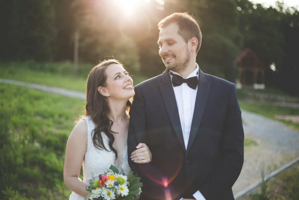 夕日の逆光の中で野の花のブーケを持ち新郎に寄り添って見上げる新婦
