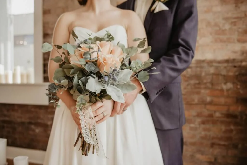 レンガの壁の前でバラとユーカリのブーケを持つ新婦と後ろに立つ新郎